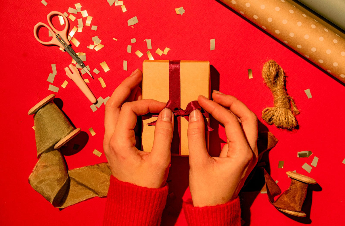 Hands tying a ribbon on a kraft gift box over a bright red background with gold confetti